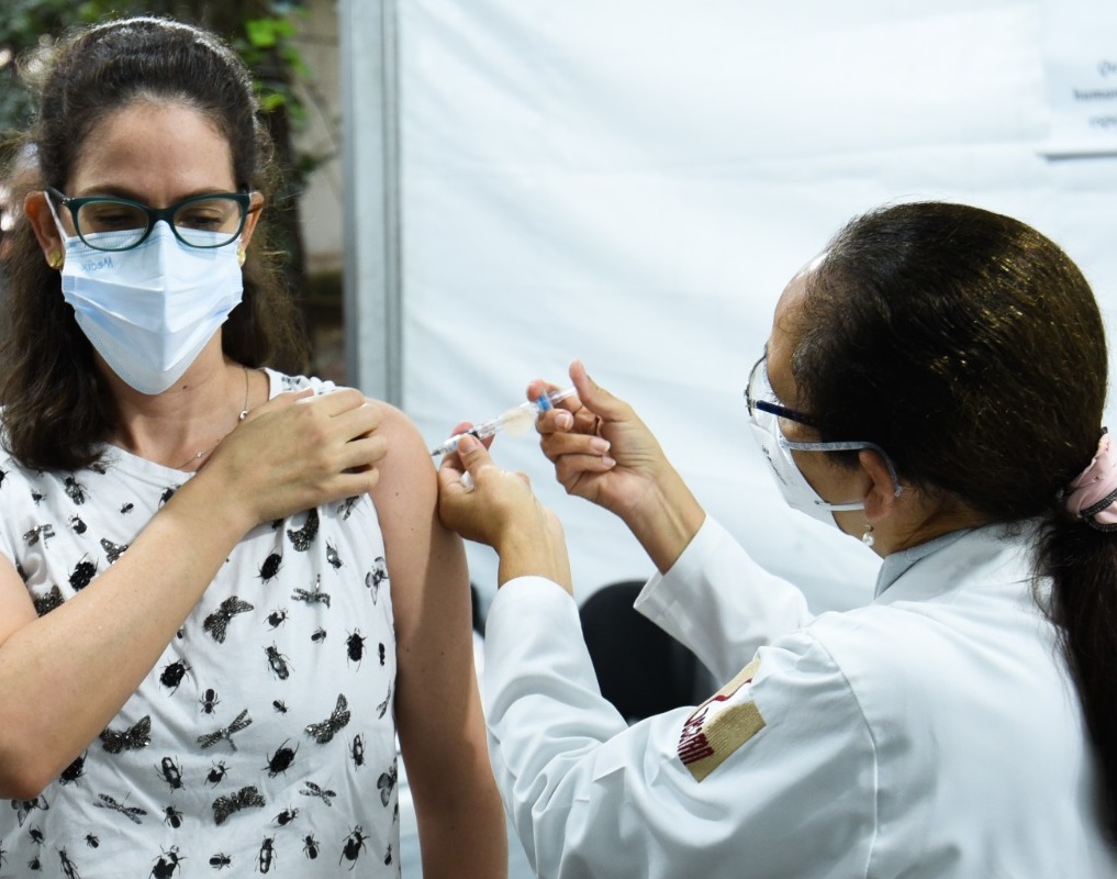 Foto de vacinação. Uma enfermeira vestindo um jaleco branco e máscara de proteção branca, com os cabelos pretos amarrados em um rabo de cavalo, presos com um acessório rosa, está aplicando uma vacina no braço de uma outra mulher. Ela está com uma das mãos levantando a manga da blusa, que é branca com borboletas pretas. A mulher está com os cabelos pretos levemente presos, usa óculos e está com uma máscara de proteção branca.